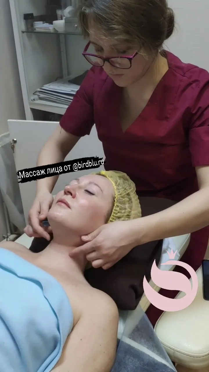 Женщина получает массаж лица в клинике, специалист в бордовом костюме проводит мягкое воздействие на область лица