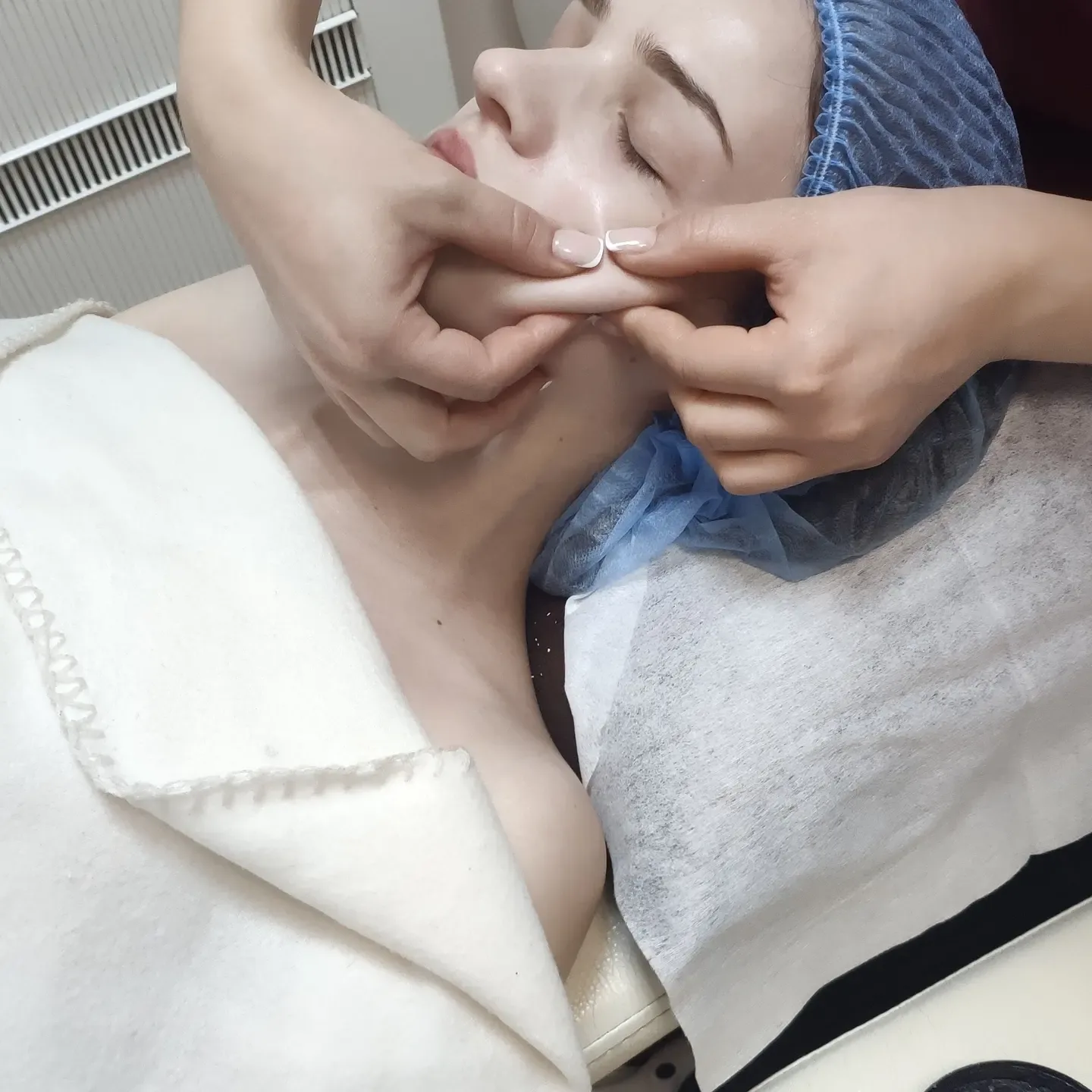 Процедура ухода за кожей лица: специалист обрабатывает зону щек у пациента в шапочке в клинике