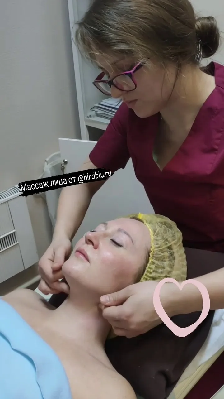 Женщина получает массаж лица в клинике, специалист в бордовой форме аккуратно поддерживает шею пациентки в шапочке
